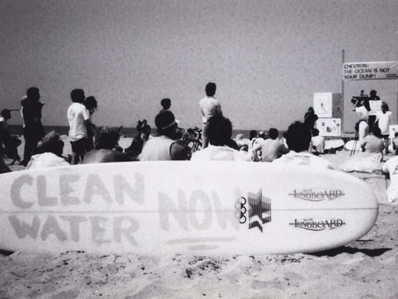 black and white image of a surfboard with the words Clean Water Now with people on the beach in the background 