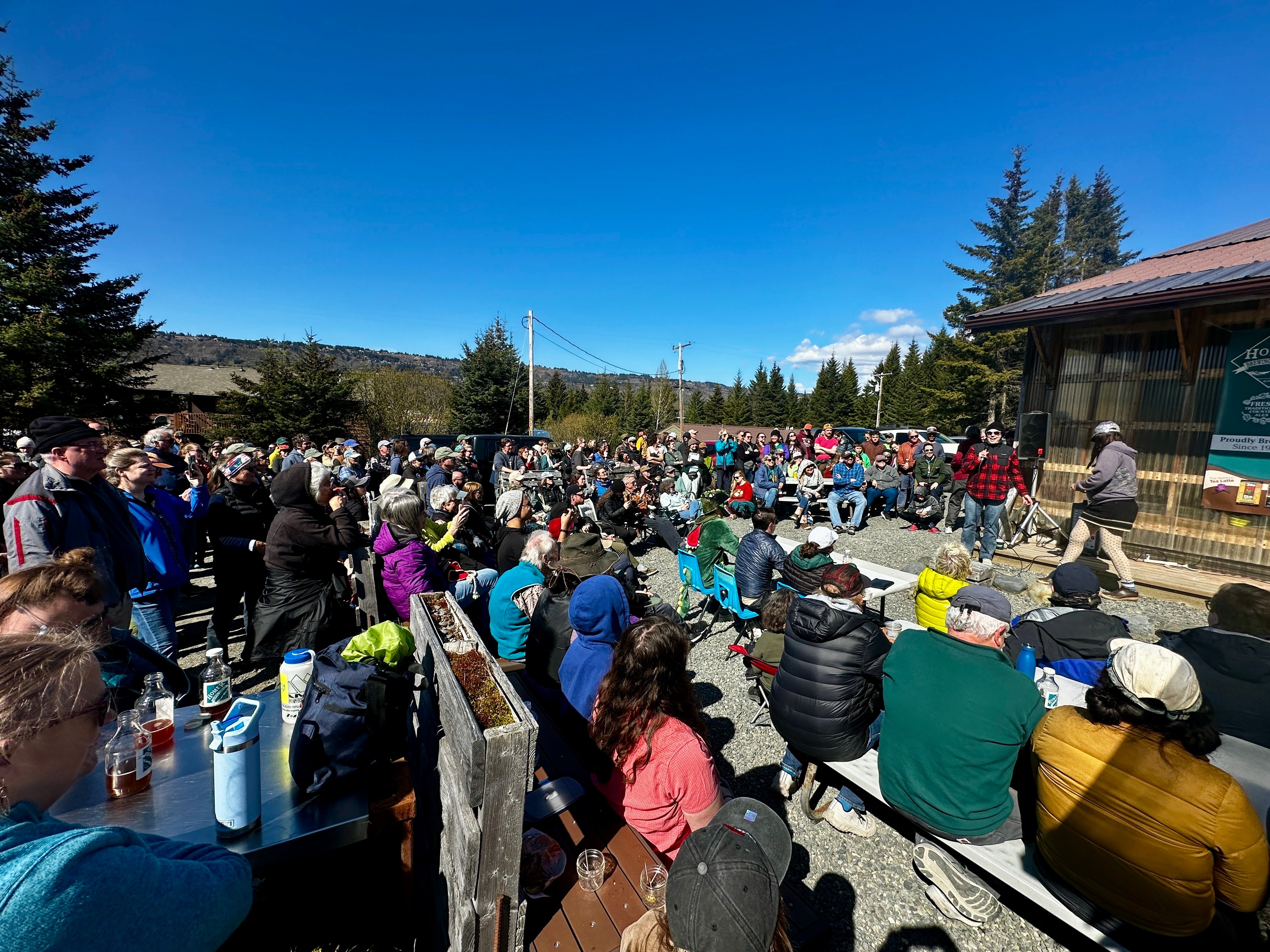 A large group of people gathered under blue skies outside, with a table with four judges observing a bird calling contest at a local brewery in Homer, AK