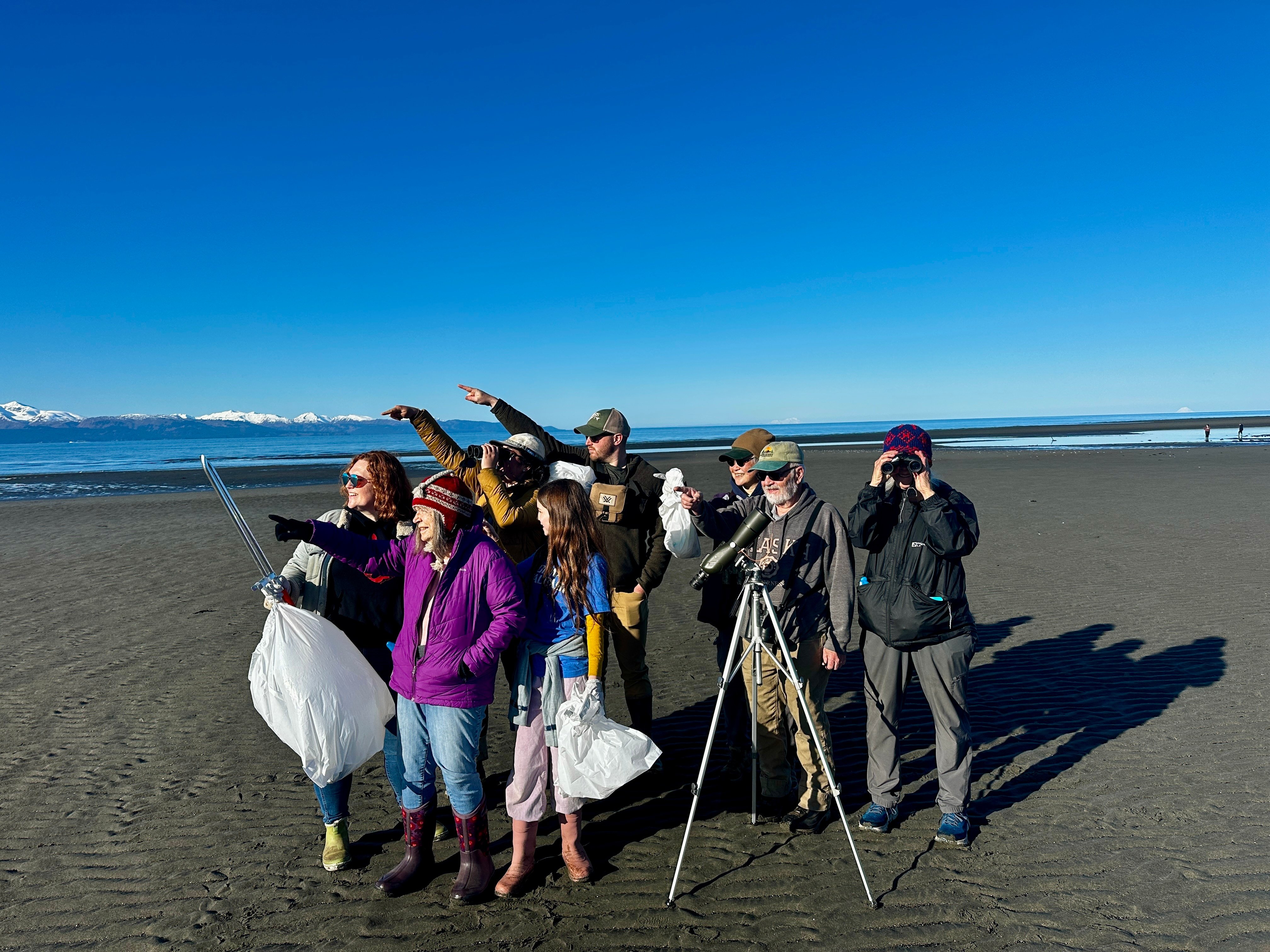 A group of people pointing into the distance, standing on the beach with binoculars, spotting scope, and trash bags, posing on a sandy beach with bright blue skies