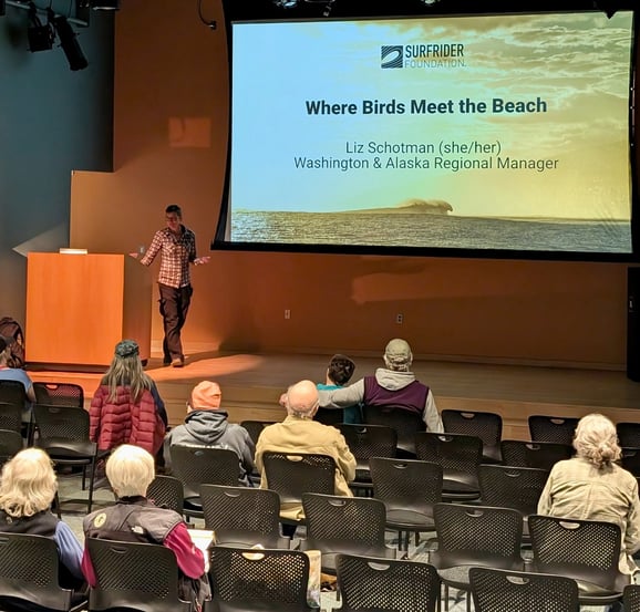 Liz Schotman stands on a stage next to a slide that reads "Where Birds Meet th Beach" with an audience watching