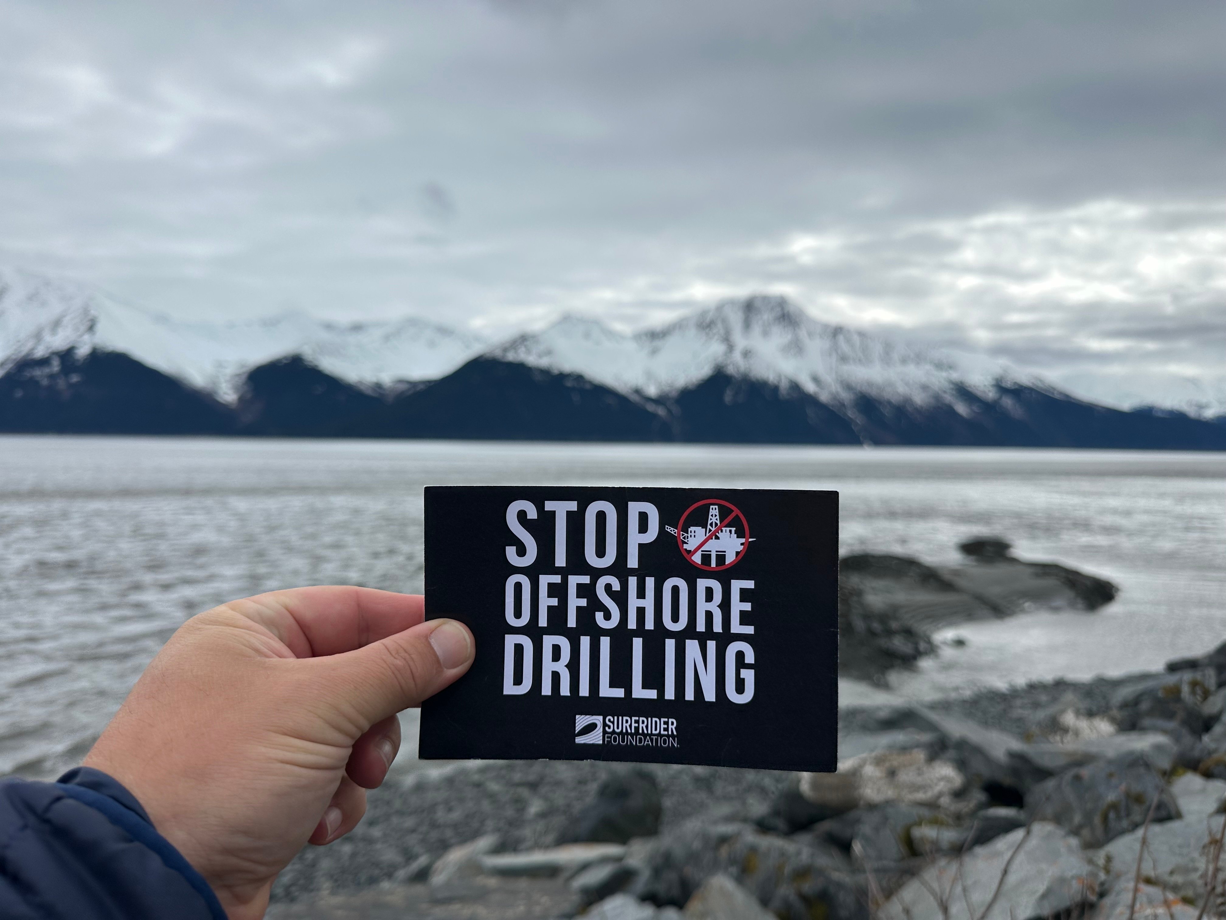 Someone holding up a Driling is Killing postcard with a snowy mountain range and rocky beach in the background