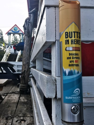 Close up shot of the blue and yellow cigarette butt canister with a dock in the background