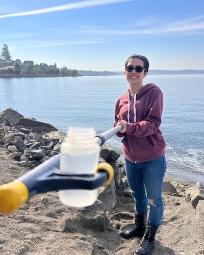 A volunteer stands on a beach on a sunny day, holding a water sample in a small bottle at the end of a boat hook 