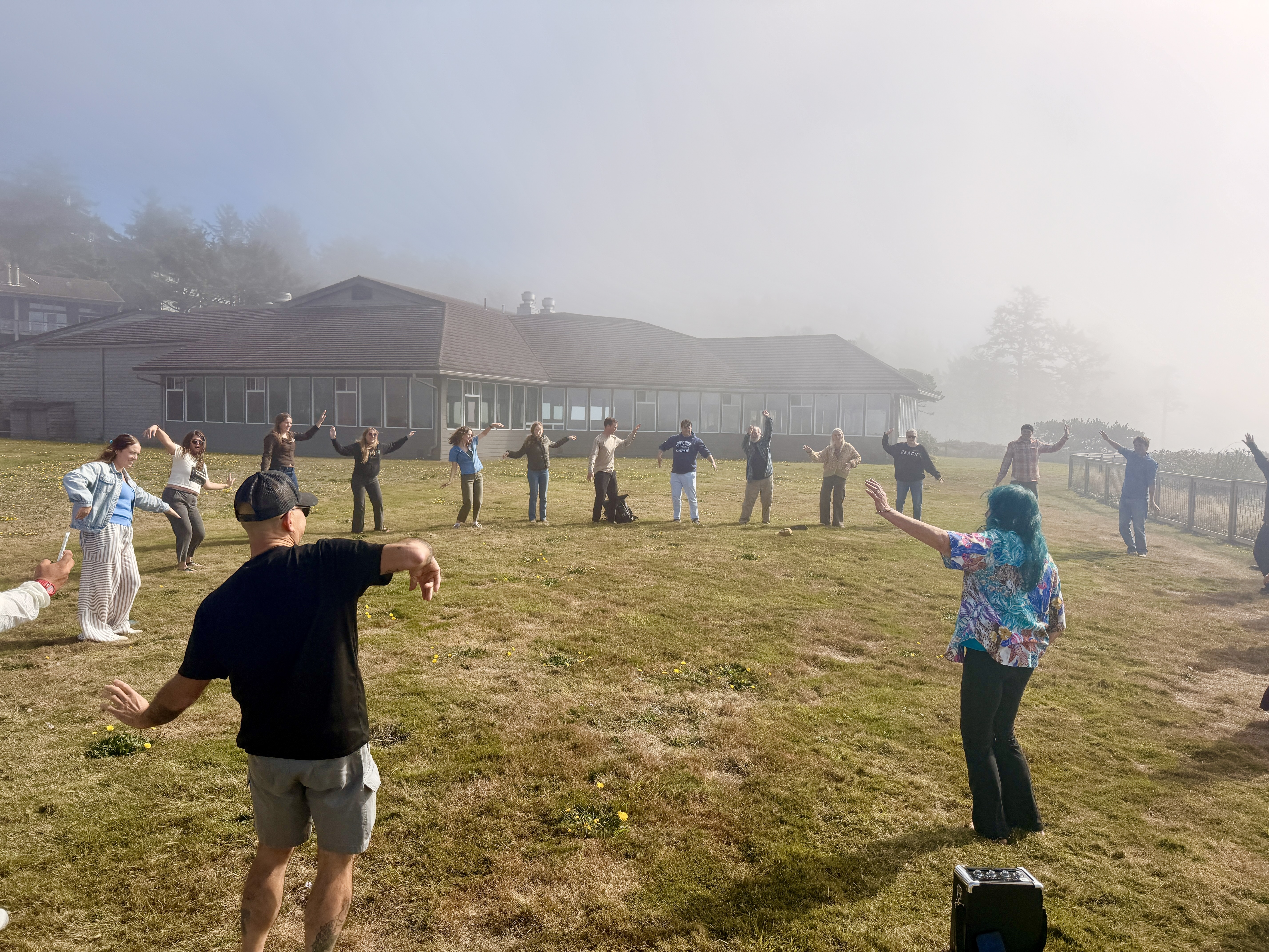 Volunteers gather in a circle outside on a misty lawn, practicing dance moves