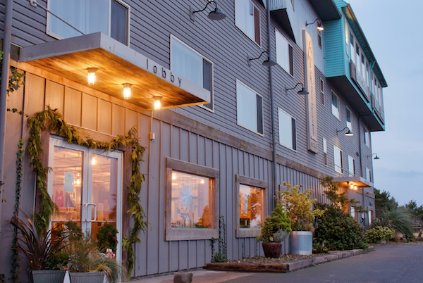 The Adrift Hotel's lobby entrance viewed from the parking lot at dusk, with warm lights illuminating the doorway and windows