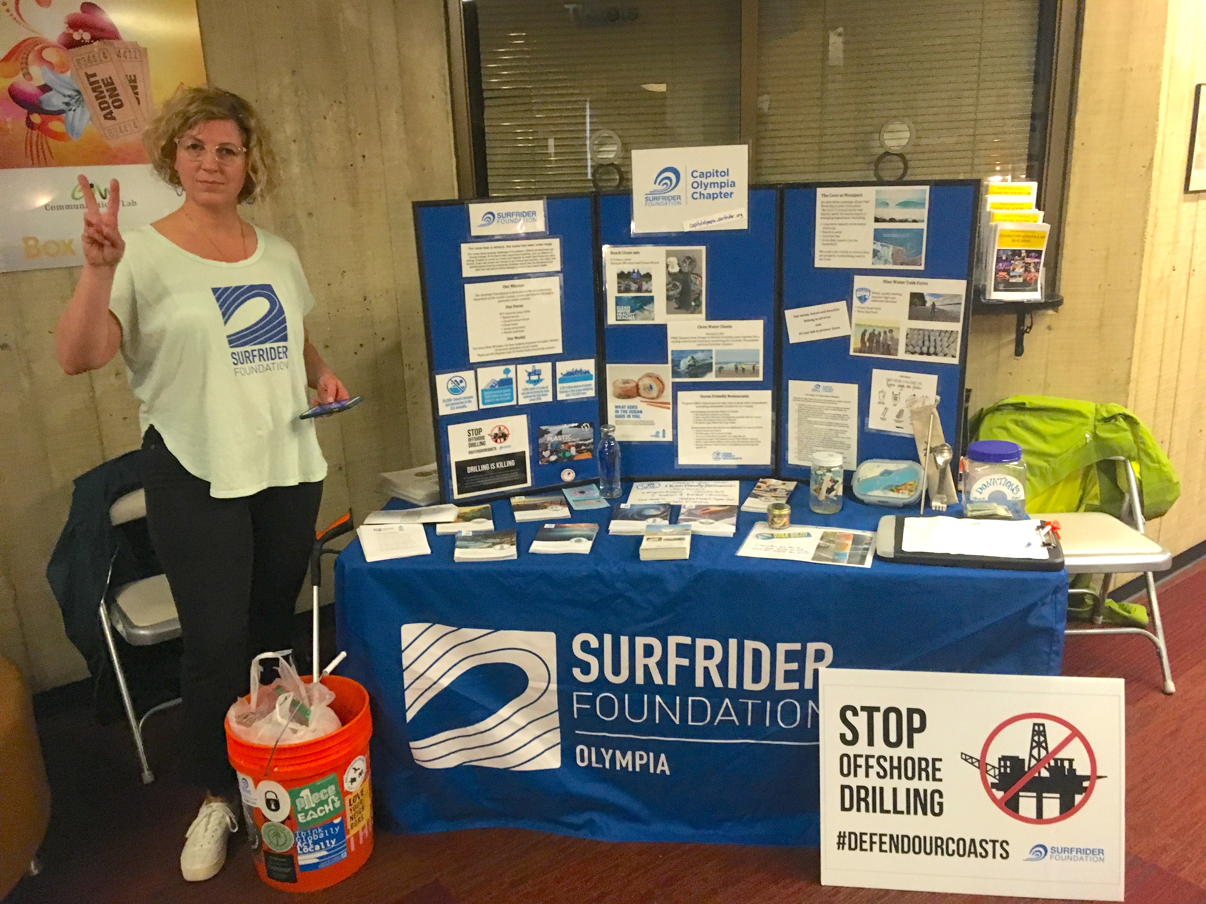 Long time volunteer Christine posing by outreach tabling materials, including a ban drilling sign and a bucket full of beach trash