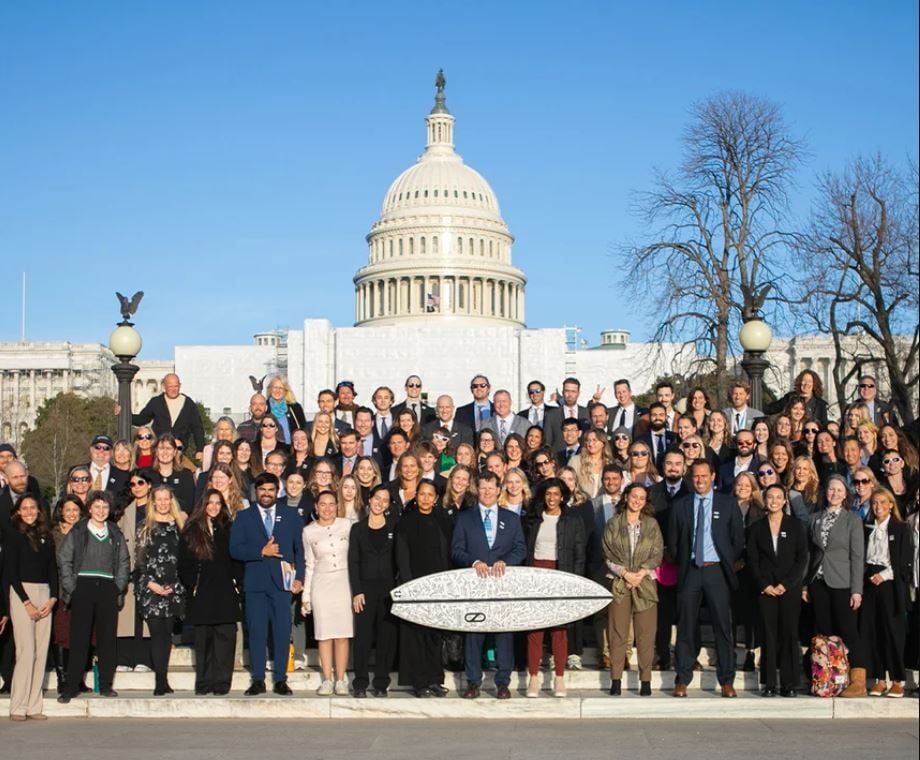Dozens of advocates pose in front of the capitol building holding a surfboard